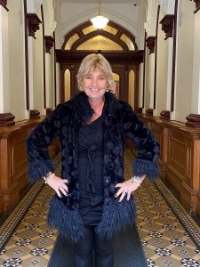 Linda is standing in a corridor wearing a black feather coat and black dress.