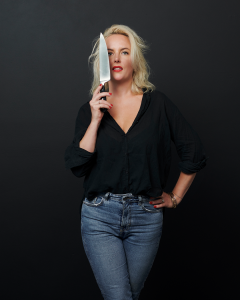 Lucy Tweed in front of a dark background, wearing jeans and a back shirt. She'd holding a chef's knife to her face.