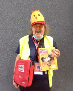 Vendor Peter smiling with the Chooks edition of The Big Issue. He has a chicken hat on his red Big Issue hat.