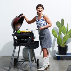 Yumi laughing at the BBQ, cooking corn. She wears big cowboy boots and wears an apron.